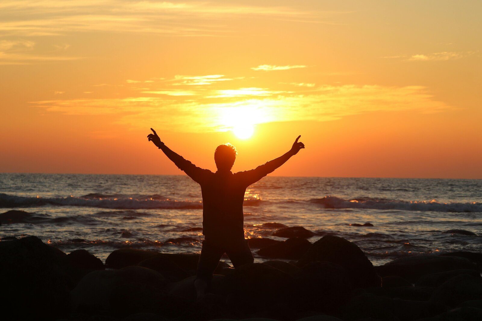 Person raising their arms beside the ocean at sunset
